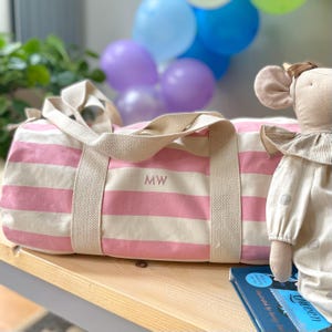 May include: A pink and white striped duffel bag with beige handles and the initials "MW" embroidered on the side. The bag is on a wooden surface, with a doll and balloons in the background. The bag is designed for travel or carrying items.
