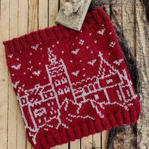 Könnte beinhalten: Ein gestrickter roter Schal mit einem weißen Schlossdesign und kleinen weißen Fledermaus- und Schneeflocken-Akzenten. Der Schal ist aus dickem Garn gefertigt und wird auf einer Holzoberfläche präsentiert. Eine kleine Gargoyle-Statue und eine Plastikspinne sind ebenfalls vorhanden.