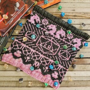 May include: A pink and black knitted dice bag with a geometric pattern and dice scattered around it. The dice are various colors and sizes.