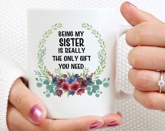 Könnte beinhalten: Weiße Keramiktasse mit einem floralen Kranzdesign und dem Text "Meine Schwester zu sein ist wirklich das einzige Geschenk, das du brauchst".