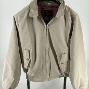 Könnte beinhalten: Eine beige Jacke mit Reißverschluss, spitzem Kragen, gerippten Bündchen und Saum sowie zwei Vordertaschen mit Knopfverschluss. Das Innere ist mit einem rot-grün-schwarzen Schottenkaro-Stoff gefüttert. Ein schwarzes Etikett mit dem Text "Cool Craft" ist sichtbar.