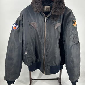 Può includere: Un giubbotto bomber nero con colletto in pelliccia sintetica marrone scuro, chiusura frontale con zip integrale, polsini e orlo a coste. Il capo presenta due inserti triangolari in pelle vicino al colletto e due toppe circolari sulle maniche.