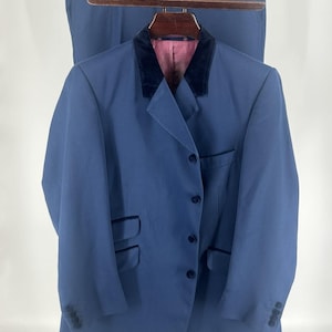 May include: A blue suit set consisting of a jacket and matching trousers. The jacket features a dark navy velvet collar, four front buttons, two flap pockets, and a chest pocket. The interior lining is a textured pink fabric with a subtle pattern.