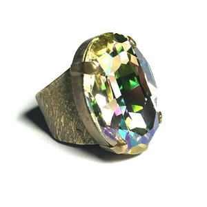 May include: A ring featuring a large, oval-shaped, iridescent crystal with multifaceted surfaces that reflect green, yellow, and purple hues. The crystal is set in a textured, dark metallic band with a rugged, uneven finish and prong settings.