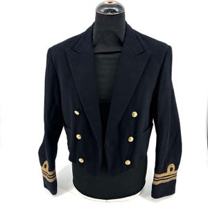 Könnte beinhalten: Eine marineblaue zweireihige Mess-Jacke mit goldfarbenen Knöpfen in zwei Reihen und goldfarbenen, nautischen Stickstreifen an jedem Ärmelabschluss. Die Jacke hat einen steigenden Reverskragen und eine kurze, taillierte Silhouette.