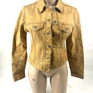 Könnte beinhalten: Eine hellbraune Wildlederjacke mit klassischem Kragen, langen Ärmeln und einer durchgehenden Knopfleiste. Das Kleidungsstück verfügt über zwei Brusttaschen mit geknöpften Klappen und vertikale Ziernähte auf der Vorderseite für eine strukturierte Passform.