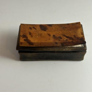 May include: A rectangular wooden box featuring a dark brown base and a lid with a lighter brown, mottled, and textured surface. The lid has a slightly curved, uneven top edge, and the overall construction shows a deep, natural wood grain finish.