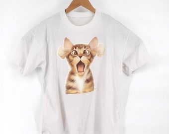 Könnte beinhalten: Ein weißes T-Shirt mit kurzen Ärmeln und einem zentralen Aufdruck einer braunen getigerten Katze mit weit aufgerissenen Augen und offenem Maul. Hinter den Ohren der Katze sind kleine weiße Wolken zu sehen, die einen humorvollen Effekt erzeugen.