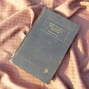 May include: A vintage, dark blue book titled "How to Be a Successful Writer" by James Irving. The book has a textured cover and is lying on a brown fabric surface.