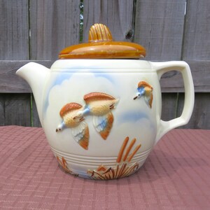 May include: Cream-colored ceramic teapot with a brown lid and handle. The teapot features a raised relief design of flying ducks, blue sky, and reeds. A vintage-style decorative piece.