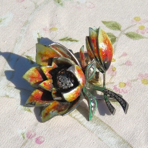 May include: Vintage floral brooch featuring a detailed flower and bud design. The brooch has a silver-tone metal base with enamel in shades of yellow, orange, and red. The flower's center is dark, and the leaves are green.