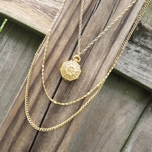 May include: A gold-colored layered necklace with a filigree pendant. The pendant is octagonal with a detailed design. The necklace chains are different styles, with one being a delicate chain and the other a thicker curb chain.