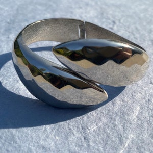 Pode incluir: Um bracelete de metal prateado com um design abstrato único. O bracelete é aberto e tem um acabamento liso e polido.