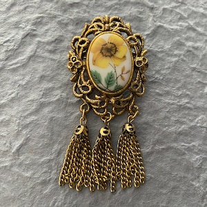 May include: Vintage gold-tone brooch. It features an oval floral centrepiece with a yellow flower on a white background. The brooch has an ornate filigree design and three dangling chain tassels, creating a classic, elegant look.