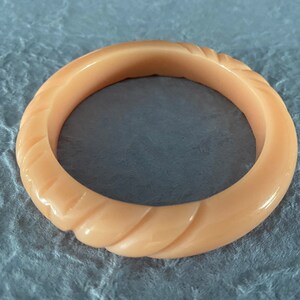 Könnte beinhalten: Ein pfirsichfarbenes Armreif-Armband mit einem geschnitzten, gedrehten Seildesign. Das Armband hat eine solide, kreisförmige Form und die Oberfläche hat ein glänzendes Finish. Der Schmuck befindet sich vor einem strukturierten, grauen Hintergrund.