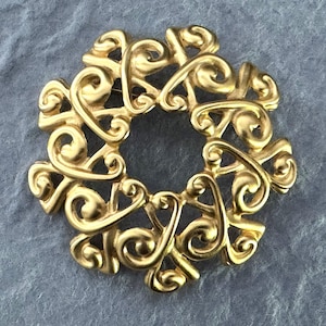 May include: A gold-toned metal brooch with an openwork design. The brooch features a circular shape with an intricate pattern of swirling, scroll-like elements. The brooch is set against a gray background.
