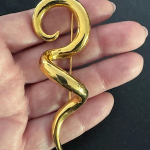 May include: A gold-toned, abstract brooch with a curved, flowing design. The pin has a long, slender shape with a spiral at the top and a pointed end. The brooch is made of shiny metal and has a smooth surface.