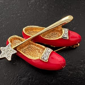 Peut inclure: Deux broches en forme de chaussures, en émail rouge et dorées. Les chaussures ont des brides et des intérieurs dorés, avec des strass sur les brides et une étoile en strass sur une chaussure. Sur fond sombre.