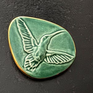 May include: A green ceramic tile with a gold rim features a raised, detailed image of a hummingbird in flight.