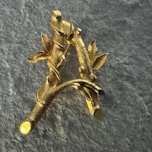 May include: A gold-coloured letter "A" shaped like a tree branch with leaves. The letter is made of metal and has a textured surface. The design is detailed and nature-inspired.