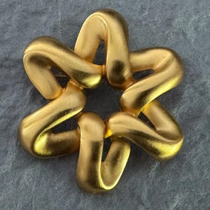 May include: A gold-toned star-shaped brooch. The brooch is made of interlocking, curved segments, creating a three-dimensional effect. The metal has a matte finish, reflecting light. The design is symmetrical and abstract.