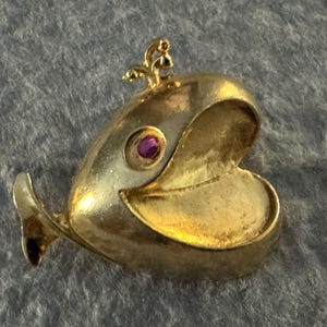 Op de afbeelding: Een gouden walvis-bedel met een roze edelsteen als oog. De bek van de walvis staat open en de bedel heeft een kleine lus aan de bovenkant om aan een ketting of armband te bevestigen.