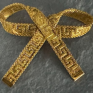 May include: A gold-colored bow-shaped accessory. The bow is made of a woven material, with a rectangular shape at the ends. The accessory is displayed on a gray surface.