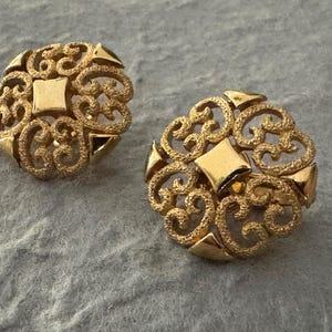 Könnte beinhalten: Ein Paar goldfarbene Ohrringe im Filigranstil. Jeder Ohrring hat eine kreisförmige Form mit einem durchbrochenen Design mit wirbelnden Mustern und einem zentralen quadratischen Element. Die Ohrringe sind vor einem grauen Hintergrund platziert.