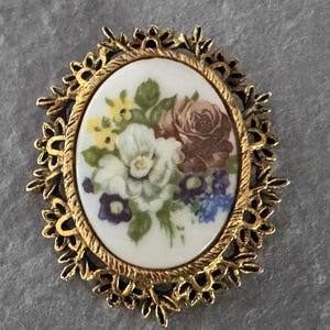 以下が含まれることがあります： 繊細な花のデザインが施された、ゴールドトーンの楕円形のブローチ。ブローチには、カラフルな花束が描かれた白い磁器のカメオが飾られています。