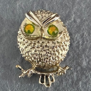 May include: Gold-toned owl brooch with textured feathers and bright yellow-green rhinestone eyes. The owl is perched on a branch, with a detailed, vintage design. A classic accessory for a jacket or scarf.