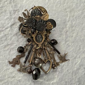 Könnte beinhalten: Eine verzierte Brosche im Vintage-Stil mit einer Mischung aus Gold- und Schwarztönen. Sie zeigt eine Ansammlung dekorativer Elemente, darunter schwarze Perlen, Schlüssel, Herzen und vogelartige Anhänger. Die Brosche hat eine detaillierte, antike Ästhetik.