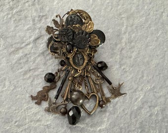May include: An ornate, vintage-style brooch with a mix of gold and black elements. It features a cluster of decorative pieces, including black beads, keys, hearts, and bird-shaped charms. The brooch has a detailed, antique aesthetic.