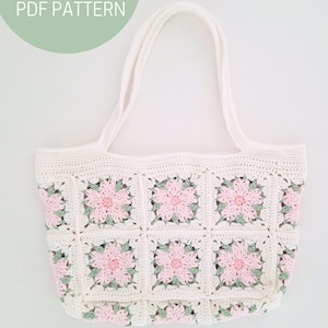 K&ouml;nnte beinhalten: Wei&szlig;e H&auml;kel-Tragetasche mit rosa und gr&uuml;nen Blumenquadraten. Die Tasche hat zwei Henkel und ist mit einem Granny-Square-Muster gearbeitet.