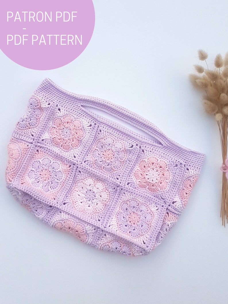 May include: A crocheted tote bag with a light purple and pink color gradient. The bag is made of square granny squares with a flower design in the center of each square. The bag has a wide opening at the top and a flat bottom.