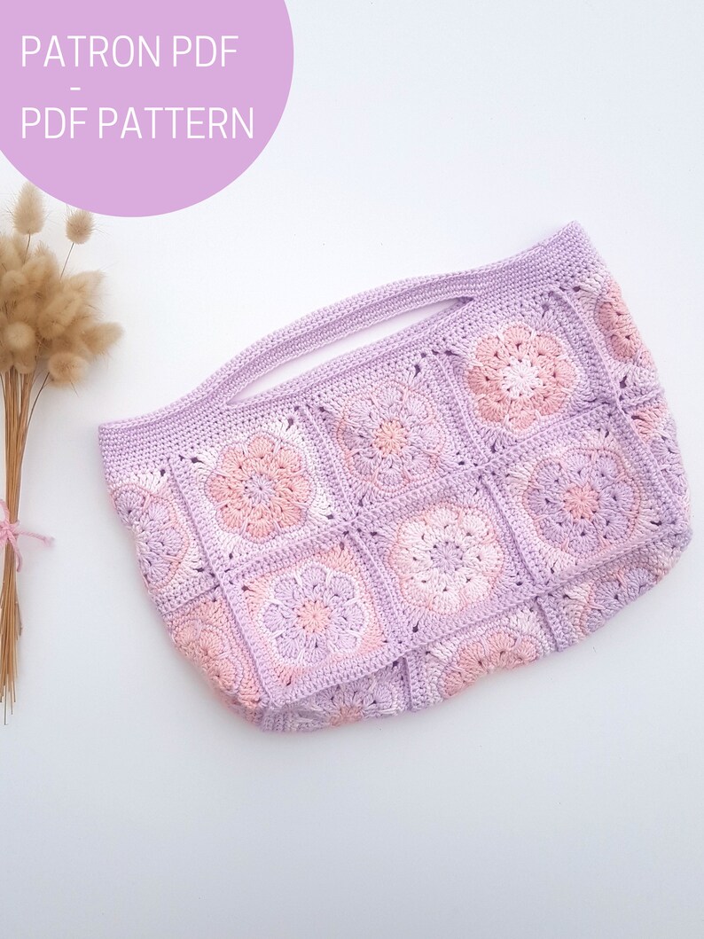 May include: A crocheted tote bag with a pink and purple floral design. The bag has a wide, flat base and two handles. The bag is made with a granny square stitch.