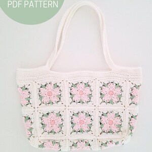 K&ouml;nnte beinhalten: Wei&szlig;e H&auml;kel-Tragetasche mit rosa und gr&uuml;nen Blumenquadraten. Die Tasche hat zwei Henkel und ist mit einem Granny-Square-Muster gearbeitet.