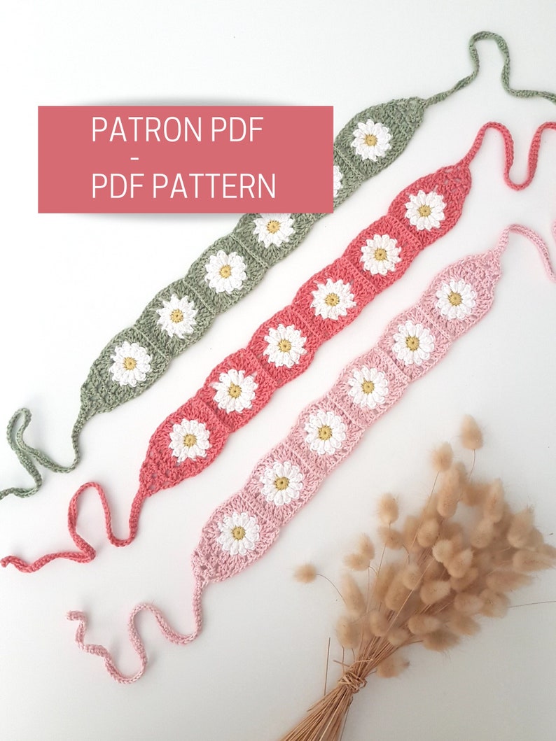 May include: Three crocheted headbands with daisy flower designs in pink, coral, and green. The headbands are laid out on a white surface with a bouquet of dried grass.