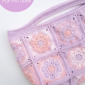 May include: A crocheted tote bag with a light purple and pink color gradient. The bag has a wide, flat handle and is made of square granny stitch motifs.