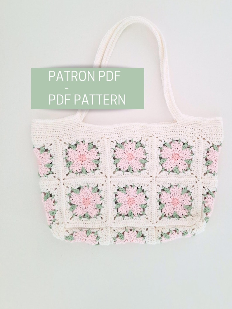 K&ouml;nnte beinhalten: Eine wei&szlig;e H&auml;kel-Tragetasche mit rosa und gr&uuml;nen floralen Akzenten. Die Tasche hat zwei Henkel und besteht aus einem geh&auml;kelten quadratischen Muster. Der Text "PATRON PDF - PDF PATTERN" ist auf dem Bild zu sehen.