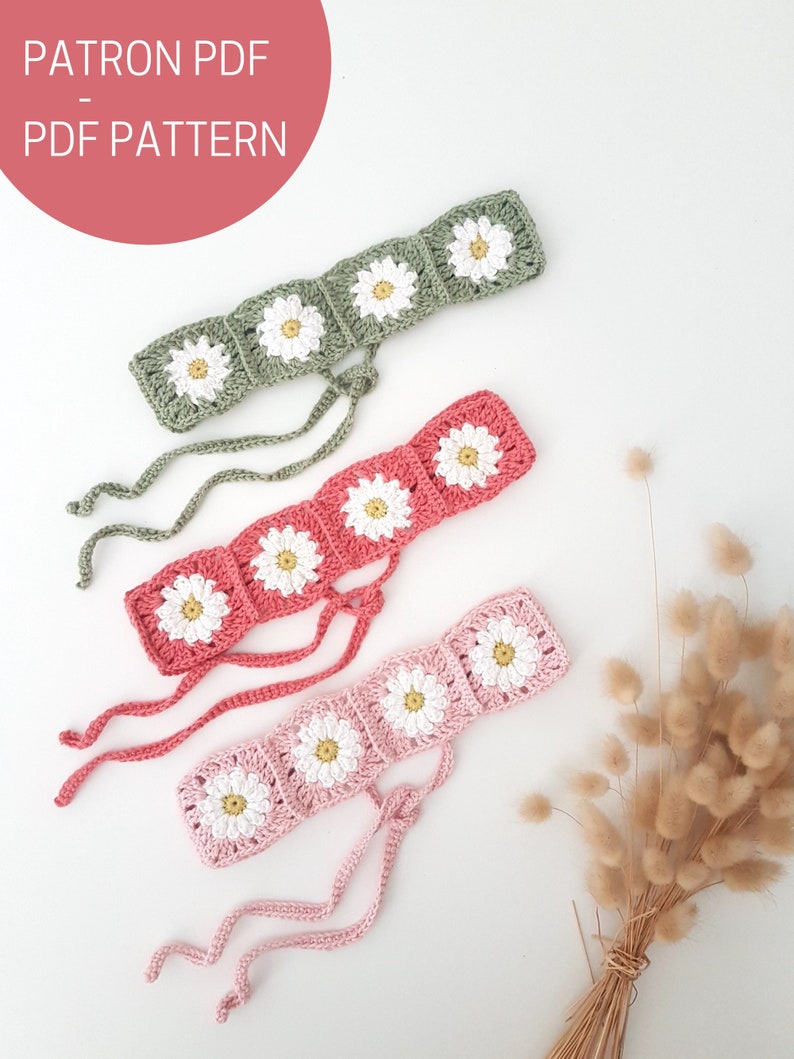May include: Three crocheted headbands with daisy flower designs in green, pink, and coral colors. The headbands are tied with long strings at the back. The headbands are displayed on a white surface with a bouquet of dried grass.