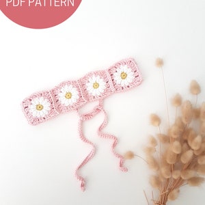 May include: A pink crocheted headband with white daisy flowers and long ties. The headband is on a white background with dried flowers.