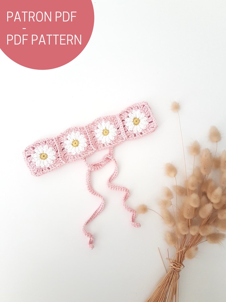 May include: A pink crocheted headband with white daisy flowers and long ties. The headband is on a white background with dried flowers.