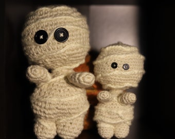 May include: Two crocheted mummy dolls with black button eyes. The larger mummy doll is standing upright, while the smaller mummy doll is sitting. Both dolls are made of white yarn.