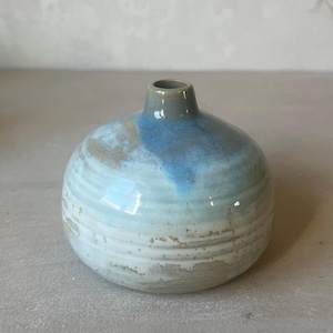Op de afbeelding: Een kleine, ronde keramische vaas met een smalle hals en een brede, bolvormige body. Het oppervlak heeft een horizontale ribbeltextuur met een glazuur dat verloopt van diepblauw aan de bovenkant naar een bleke, gevlekte witte en grijze afwerking aan de basis.