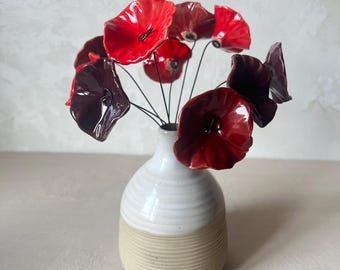 May include: A ceramic vase with a white glazed top and a ribbed beige base, containing several glossy red and dark maroon ceramic flowers attached to thin black wire stems that extend outward from the narrow neck of the vessel.