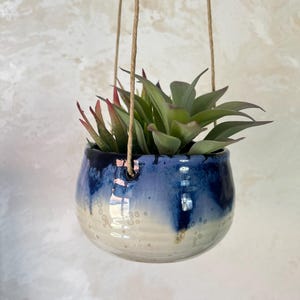 Op de afbeelding: Een ronde hangende keramische plantenpot met een glanzend glazuur dat verloopt van diep marineblauw aan de bovenkant naar een romig gebroken wit aan de basis. De pot heeft twee gaten om op te hangen en bevat een groene vetplant met rode bladpunten.