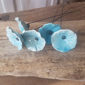 Op de afbeelding: Vijf blauwe keramische bloemen met zwarte centra op draadstengels. De bloemen zijn gerangschikt op een stuk verweerd hout.