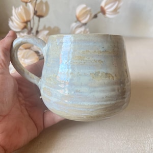 Peut inclure: Une tasse en céramique avec un corps arrondi et une anse incurvée. La surface présente un émail moucheté dans des tons bleu pâle, crème et beige terreux, avec des stries horizontales visibles sur la paroi extérieure et une finition lisse.