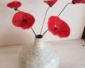 May include: A white ceramic vase with a speckled pattern holds several red ceramic poppy flowers. The flowers have black stems and centers. The vase is round and sits on a white surface.