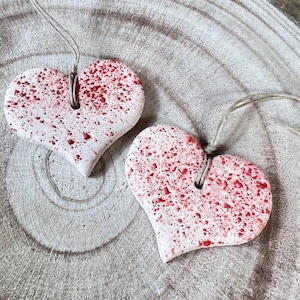 Könnte beinhalten: Zwei herzförmige Ornamente mit einem gesprenkelten roten Design auf weißem Grund. Jedes Ornament hat ein Loch oben mit einer Schnur zum Aufhängen. Sie werden auf einer hellbraunen, strukturierten Holzoberfläche präsentiert.
